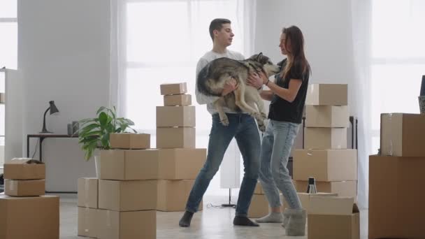 Jong getrouwd stel met schattige hond hasti dansen van vreugde in hun nieuwe appartement. Er zijn veel bewegende kartonnen dozen in de kamer.. — Stockvideo