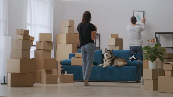 Egy férfi felakaszt egy képet a falra. A barátnő áll a távolban, és segít a szint a horizont. új élet kezdete — Stock videók