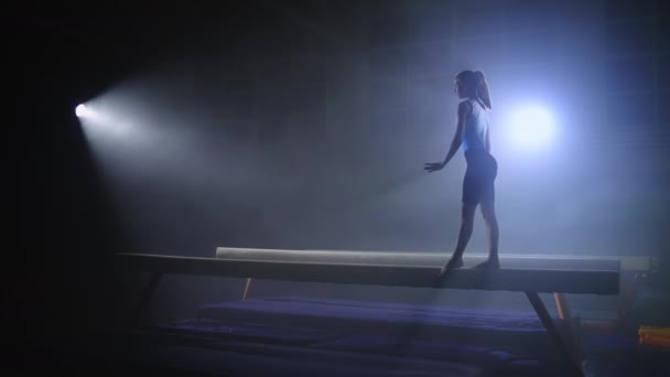 겨우살이를 하고 있는 십 대 운동 선수 소녀는 밤에 체조 홀에서 균형 광선을 넘어 어두운 스포츠 홀에서 혼자 훈련을 받고 있다 — 비디오