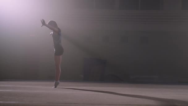 Sportief meisje is het uitvoeren van back flip op de vloer in gymnastiek hal, jonge sporter is de opleiding alleen in donkere hal — Stockvideo