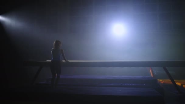 Teen girl is performing tricks on balance beam in gym hall, training and preparing to competition, silhouette of figure — Stock Video