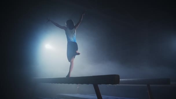 Young female gymnast is training on balance beam in dark sports hall, children sport school, rear view — Stock Video