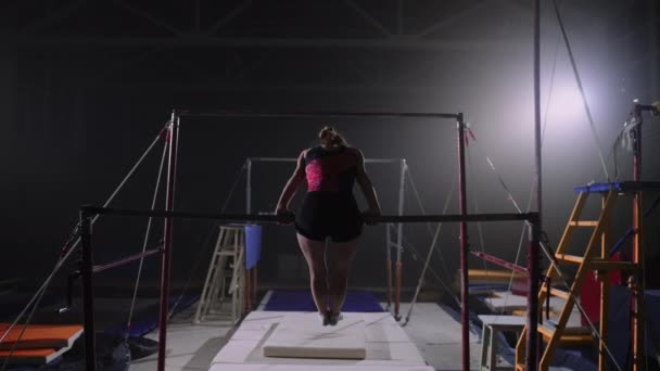 Ung idrottskvinna tränar på ojämna barer i gymnastiksal på kvällen, sportig person förbereder sig för tävling — Stockvideo