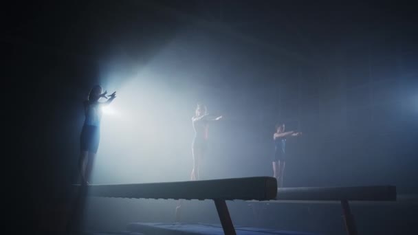 세 명의 여자 체조 선수들이 어두운 체조 홀에서 예술 체조 선수들로 이루어진 팀을 이루어 균형을 잡는 일을 하고 있다 — 비디오