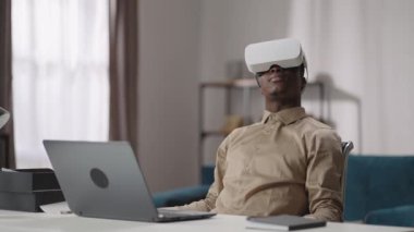 Genç siyahi adam video oyunları oynamak için kafaya monte edilmiş ekranı kullanıyor, masada dizüstü bilgisayarla oturuyor ve etrafa bakıyor.