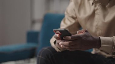 Genç siyahi adam evde akıllı telefon kullanıyor, internet sitelerine bakıyor, sosyal ağlara bakıyor, ellere yakından bakıyor.