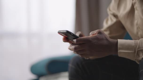 Fiatal afro-amerikai férfi szörfözik az interneten okostelefonnal, olvasás és üzenetek küldése a szociális hálók, ül egyedül otthon — Stock videók