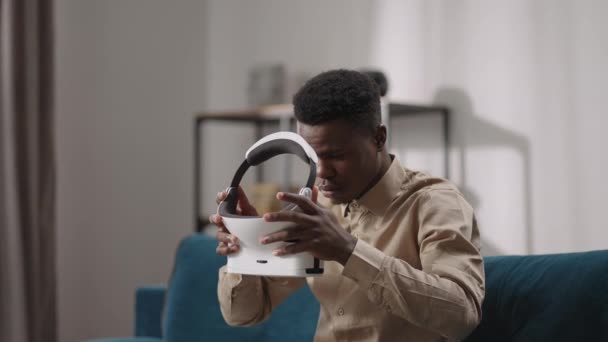 Hombre negro joven está poniendo la pantalla montada en la cabeza en la cabeza, sentado solo en la habitación, realidad virtual, descansando en casa — Vídeo de stock