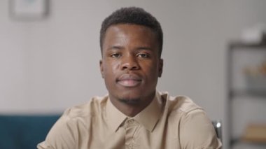 Genç Afro-Amerikalı adam kameraya bakıyor ve gülümsüyor. Dostane bir gülümseme, dairede yakın plan bir portre, siyah adamın yakın plan portresi.