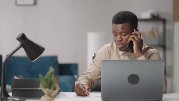 Adult black man is communicating by mobile phone, sitting at table with laptop, writing notes, working remotely from home, portrait in living room — Stock Video