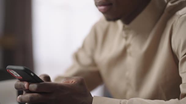 Ung afro-amerikansk man skickar meddelanden via smartphone, sitter vid bordet med bärbar dator, närbild från händer till ansikte — Stockvideo