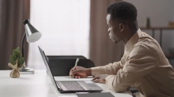 Inteligente estudante negro do sexo masculino está fazendo anotações, sentado na frente do laptop, estudando em casa, trabalho à distância e educação — Vídeo de Stock