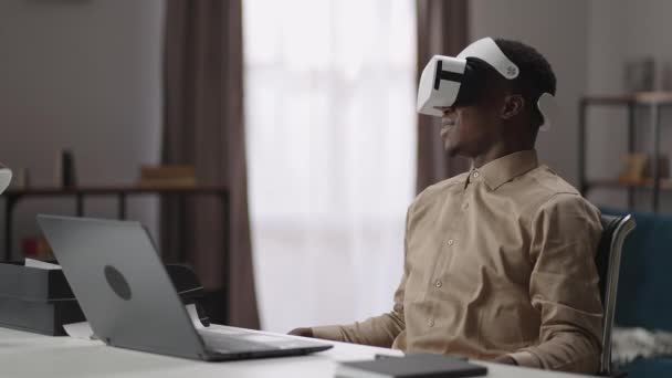 Νεαρός μαύρος άνδρας χρησιμοποιεί γυαλιά VR, κάθεται στο σπίτι, στο τραπέζι με ανοιχτό φορητό υπολογιστή, Αφρο-Αμερικανός τύπος βλέπει γύρω — Αρχείο Βίντεο