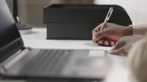 Afro-americká osoba píše poznámky na papír, sedí u stolu s notebookem, studuje online, detailní záběr rukou — Stock video