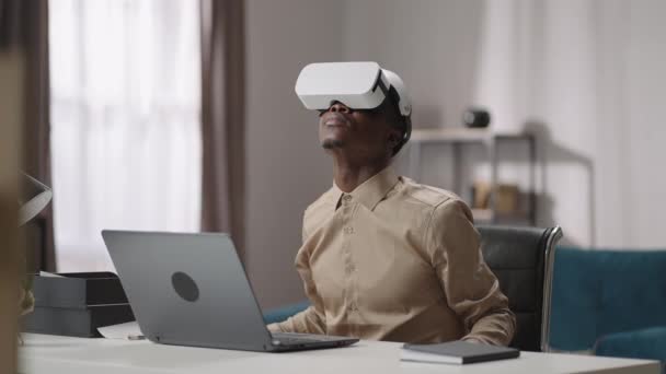 Usando la realidad virtual para la educación, el joven negro está usando la pantalla montada en la cabeza, sentado en la mesa con el ordenador portátil y mirando a su alrededor — Vídeo de stock