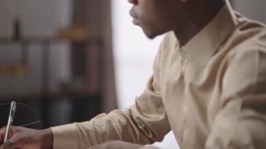 Genç siyahi erkek yazar veya gazeteci evde çalışıyor, kağıt üzerine kaba taslak yazıyor, dizüstü bilgisayarla masada oturuyor