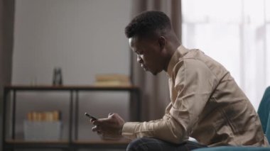 Afro-Amerikalı öğrenci akıllı telefon kullanıyor, evde tek başına oturuyor, sosyal ağlarla iletişim kuruyor, erkek alet kullanıcısı.