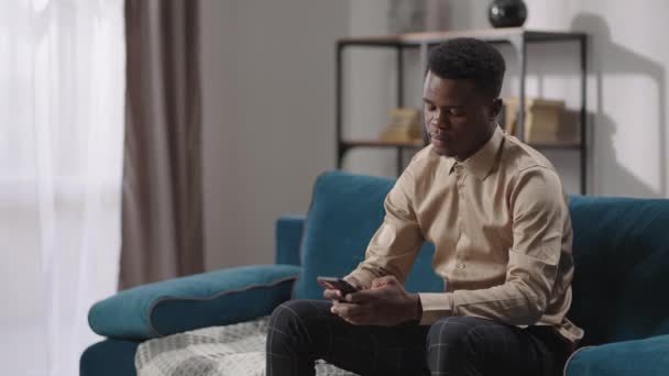 Νεαρός Αφρο-Αμερικανός άνθρωπος χρησιμοποιεί smartphone κάθεται στον καναπέ στο διαμέρισμα, αρσενικό χρήστη του Διαδικτύου, προβολή κοινωνικών δικτύων — Αρχείο Βίντεο