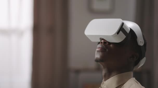 Афро-американський хлопець використовує сучасний дисплей на голові в кімнаті, сидить і озирається навколо, насолоджуючись віртуальною реальністю — стокове відео