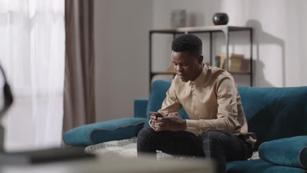 Afro-Amerikalı genç akıllı telefon kullanıyor, evde tek başına oturuyor, genç siyah adam mesaj yazıyor ve sosyal ağlara bakıyor. — Stok video