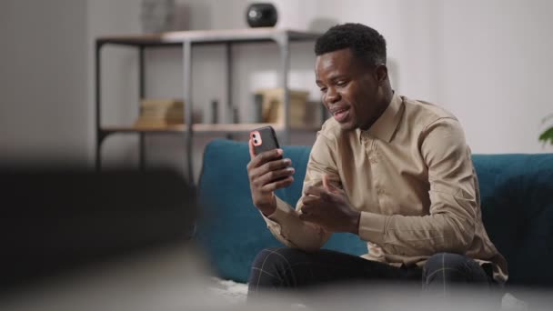 ชายแอฟริกัน-อเมริกันที่มีความสุข กําลังคุยกันทางวิดีโอโทรศัพท์ในสมาร์ทโฟน นั่งคนเดียวบนโซฟา ในห้องนั่งเล่นของอพาร์ทเม้นท์ วิดีโอแชทกับเพื่อนในเครือข่ายสังคม — วีดีโอสต็อก