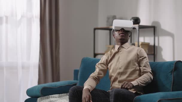 Junger afroamerikanischer Mann mit modernem Head-Mounted-Display schaut sich um, sitzt auf der Couch in der Wohnung, Technologie für Unterhaltung und Bildung — Stockvideo