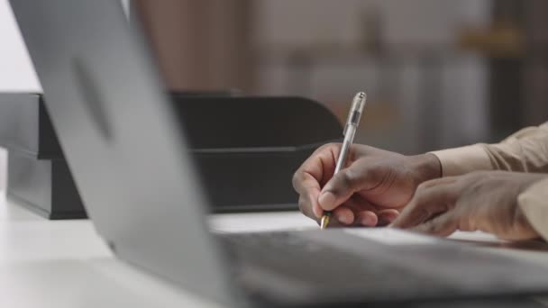 Czarny mężczyzna pisze na papierze, siedzi przy stole w jego biurze pracy, widok z bliska na ręce, laptop na pierwszym planie, młody biznesmen pracuje — Wideo stockowe