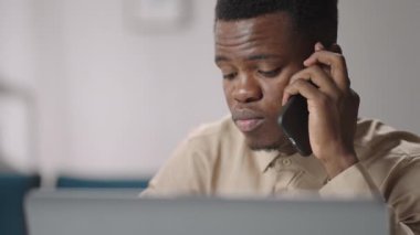 Siyah adam cep telefonuyla iletişim kuruyor, evde oturuyor ve laptopla çalışıyor, Afro-Amerikan portresini yakından çekiyor.