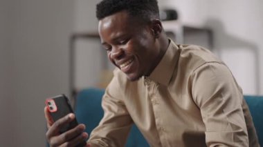 Neşeli siyahi adam akıllı telefonuyla konuşuyor, evde tek başına kanepede oturuyor, orta boy portre.