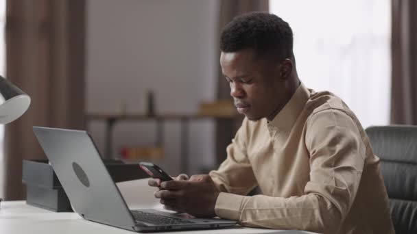 Czarny mężczyzna pracownik biurowy lub freelancer jest za pomocą smartfona podczas pracy z laptopem, siedzi przy stole, surfing internet — Wideo stockowe