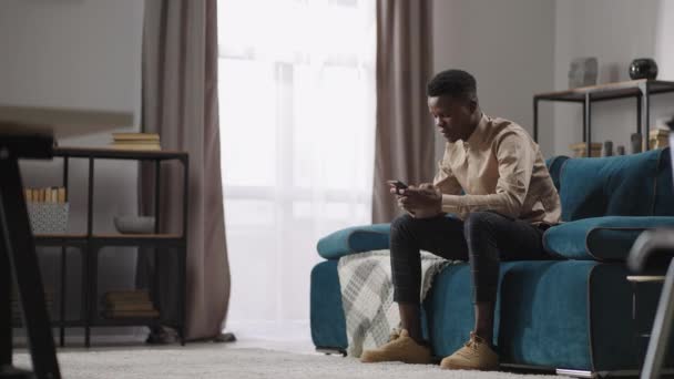 Afro-american man använder smartphone för kommunikation och spela spel, sitter hemma ensam, svart kille skriver meddelande och rulla skärmen — Stockvideo