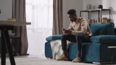 Genç siyahi adam akıllı telefonuyla video oyunu oynuyor, evde tek başına oturuyor, Afro-Amerikalı adam cep telefonuyla sosyal ağlarla iletişim kuruyor.