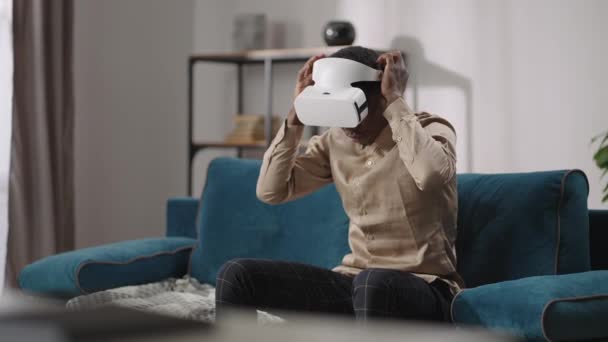 Αφρο-Αμερικανός έφηβος αγόρι βάζει στο κεφάλι-τοποθετημένα οθόνη, κάθεται στον καναπέ στο διαμέρισμα, προβολή εικόνας ή βίντεο στην εικονική πραγματικότητα — Αρχείο Βίντεο