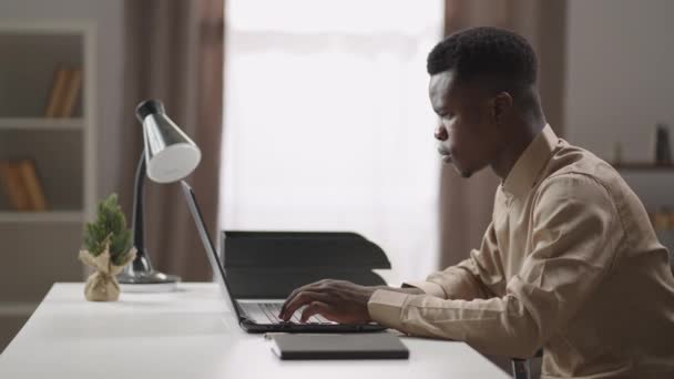 Νεαρός μαύρος άνδρας εργάζεται εξ αποστάσεως από το σπίτι, ελεύθερος επαγγελματίας σερφάρει στο διαδίκτυο στο σημειωματάριο, κοιτάζοντας στην οθόνη — Αρχείο Βίντεο
