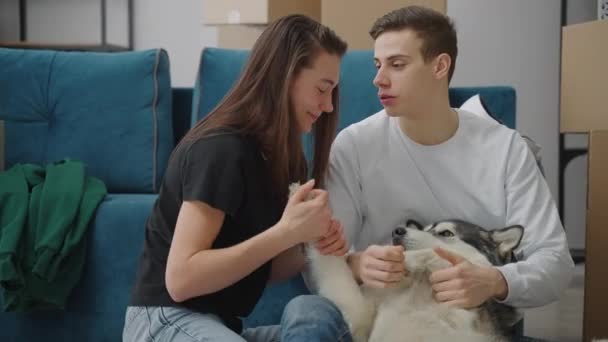 Cheerful young couple playing with their dog indoors. The man strokes the dogs face, and the woman touches the paws and stomach. The guy tells the girl a funny story. — Stockvideo