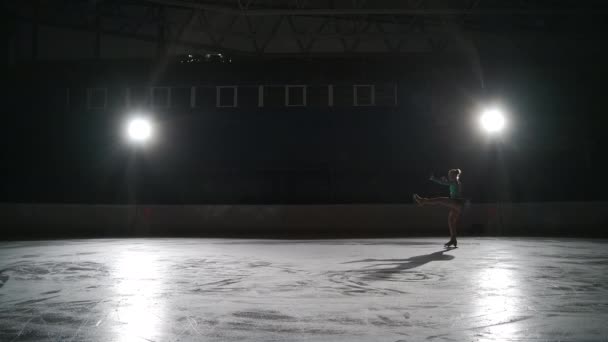 Дівчина-підліток готується до змагань з фігурного катання, юнацька спортсменка катається на ковзанах на темному стадіоні, рухаючись проти яскравого софіту — стокове відео