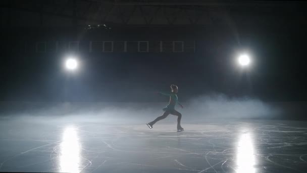 Молодая фигуристка выступает на катке в темноте, маленькая фигурка движется против яркого soffit внутри драматического тумана — стоковое видео