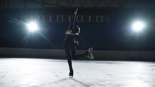 Pełna wdzięku kobieta łyżwiarka figurowa wykonuje obrót na lodowisku w ciemności, poruszając się przeciwko jasnej strofie, sylwetka — Wideo stockowe