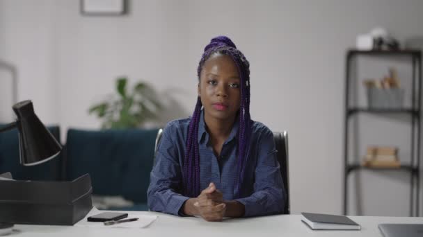 Счастливая молодая афроамериканка сидит за столом в своем домашнем офисе и смотрит в камеру, улыбаясь дружелюбно, в онлайн чате. — стоковое видео