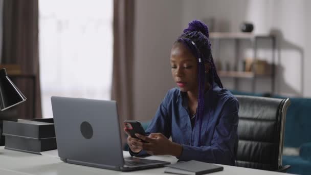 Giovane donna di colore è seduto nel suo posto di lavoro a casa e utilizzando smartphone, la visualizzazione di notizie nastro nelle reti sociali, giocare — Video Stock