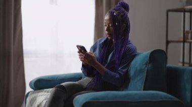Modern akıllı telefonlu afro-amerikan bayan evde tek başına oturuyor, kanepede oturuyor ve sosyal ağlarda haber okuyor, oyunlar oynuyor.