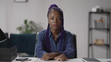 Ciddi Afro-Amerikan kadın portresi, masada oturuyor ve kameraya bakıyor, arkadaşça gülümsüyor, online sohbet kavramı.
