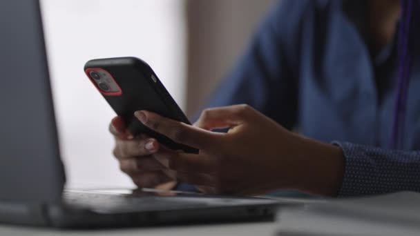 젊은 흑인 여성은 사무실에서 노트북을 들고 테이블에 앉아 메시지와 이메일을 확인하고 현대 스마트폰을 사용하고 있습니다. — 비디오