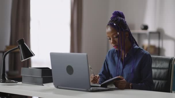 Genç siyahi iş kadını, ev ofisinde çalışıyor, dizüstü bilgisayarla masada oturuyor, not defterine yazıyor ve planlıyor. — Stok video