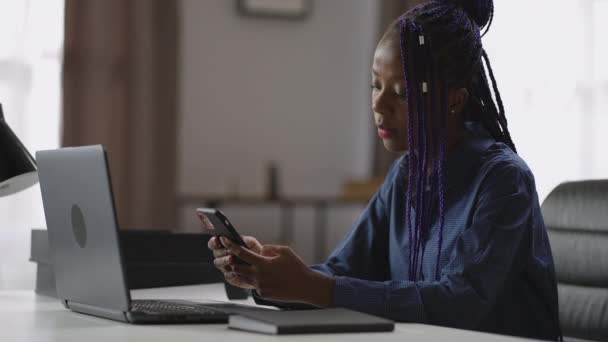 Afro-Amerikaanse vrouwelijke werknemer rust in haar kantoor tijdens de werkdag, met behulp van smartphone, het controleren van e-mail en chatten in sociale netten — Stockvideo