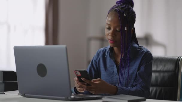 美国黑人女士在上班时间使用智能手机，在社交网络上查看邮件和信息，带着笔记本电脑坐在桌旁 — 图库视频影像