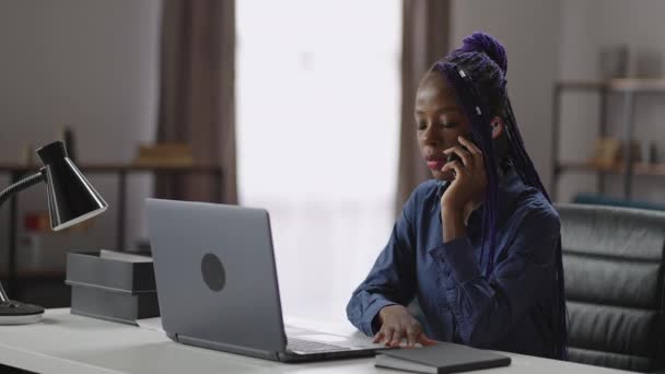 Czarna kobieta dzwoni przez telefon komórkowy, siedzi przy roboczym stole w swoim biurze, rozmawia przez telefon komórkowy, średni portret — Wideo stockowe