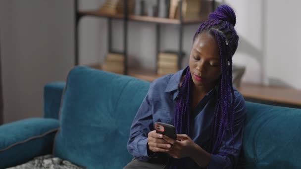 在现代公寓的沙发上，身穿智能手机的年轻黑人妇女的肖像，非洲裔美国女士正在用手机上网 — 图库视频影像