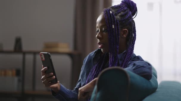 흑인 여성은 그녀의 아파트 거실에 앉아서 스마트폰으로 일하는 비디오 채팅을 듣고 스크린을 보고 있습니다. — 비디오