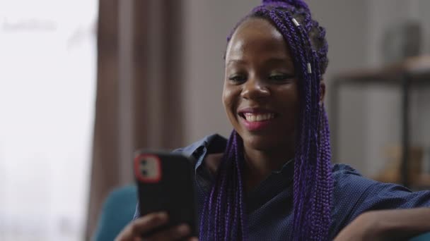 쾌활 한 흑인 여성 이 스마트폰으로 비디오 통화를 하며 크게 웃고, 진열대를 보며 웃고, 집에서 쉬고 있습니다. — 비디오